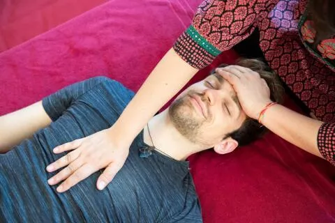 A practitioner places hands on a relaxed man's forehead and chest during an energy healing session.